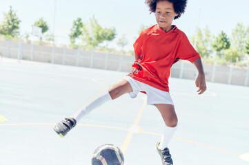 Young afro American boy with red football shirt playing football in a training field. Learn to dribble.