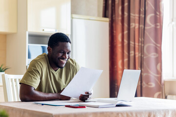 latin hispanic man looking happy while sitting with laptop and papers at kitchen