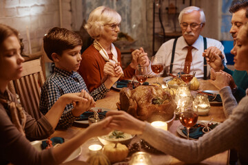 Small boy and his extended family holding hands while praying before Thanksgiving dinner at dining table.