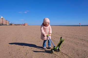 Little girl in a coat dragging her scooter through the sand on an empty beach