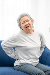 senior woman sitting on a sofa at home with a backache, feeling pain and with an expression of being unwell. Health problem concept.