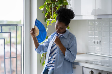 Exhausted African American woman standing in kitchen holding paper fan and feeling unwell due to...