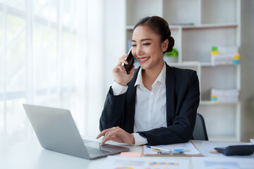 Beautiful Asian business woman chatting on the phone with customer and working on her laptop and...