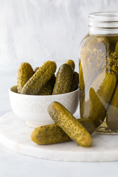 A bowl and jar of dill pickles with two pickles in front on a marble slab.