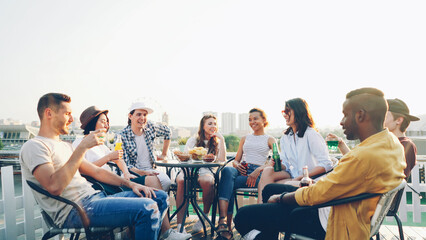 Multiracial group of friends is having open air party drinking, talking and laughing enjoying summer and good company. Youth, big city and celebration concept.