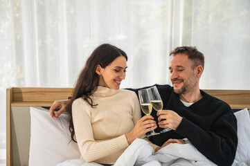 European couple smiling in winter clothing holding glasses of champagne sitting on bed under blanket.