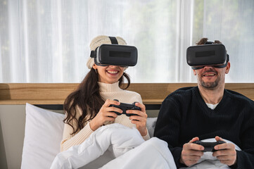 Caucasian couple wearing a VR headsets goggle device and holding game controller sitting on their bed playing game together.