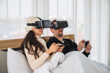 Caucasian couple wearing a VR headsets goggle device and holding game controller sitting on their bed playing game together.