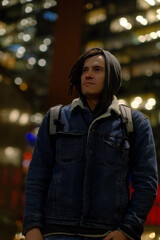 Portrait of young man with dreadlocks under street lamp at night on background of high-rise buildings with lighting from windows.