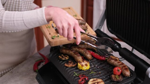 Woman putting Grilled Homemade juicy pork sausages from electric grill to serving board