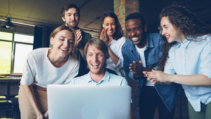 Excited young man is looking at laptop screen, rejoicing and expressing happiness, his happy colleagues are congratulating him on success, clapping hands