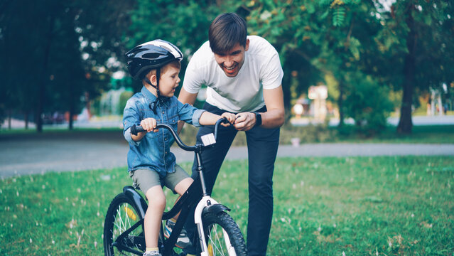 Handsome young man loving father is teaching his small son to ride bicycle in park on summer day, cute little boy is riding bike while dad is holding him and running.