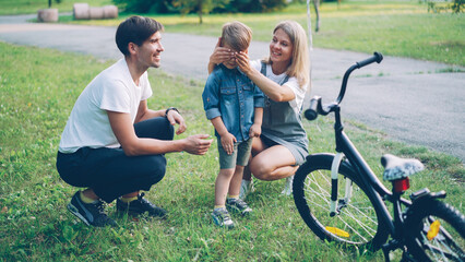 Loving parents are making surprise for little son closing his eyes and giving him new bicycle as present, happy excited boy is looking at bike and talking to mother and father.
