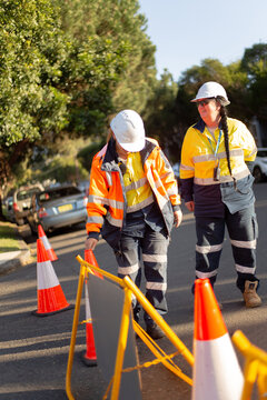 Two women road workers with white helmet orange and yellow jackets on the road with cones