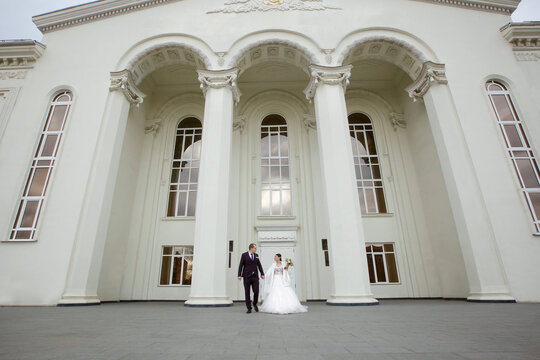 Happy loving wedding couple on a walk. Bride in white dress and veil. The groom in a jacket. In the background are white pillars.