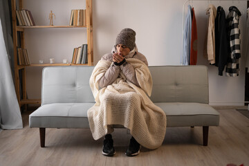 Unwell man in hat and blanket sit in cold living room suffer from heat lack. Unhealthy young man...