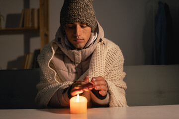 A man dressed in a warm winter hat sits at home at a table and warms his hands from a burning...