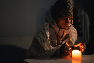 A young man dressed in a warm winter hat at home at a table and warms his hands from a burning...