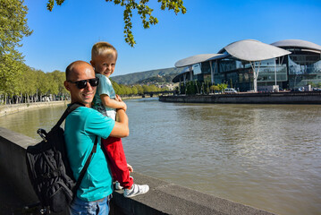 Tbilisi Georgia April 27 2019. A young man with a child on the embankment of the Kura river Mtkvari with the Tbilisi public service hall located on the Bank in the heart of the city.