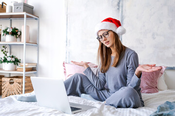 Emotional girl sitting at home with laptop in santa hat gesticulates with her hands in happiness. Festive mood of winter holidays of christmas and new year