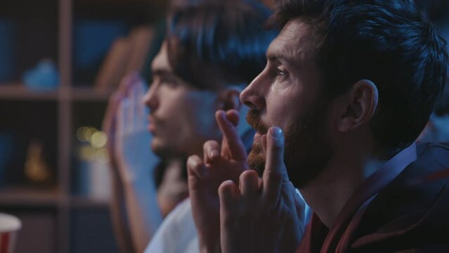 Millennial people sitting on couch at home and awaiting for favorite football team victory. Nervous friends crossed fingers with concentrated face expressions and praying for the winning.