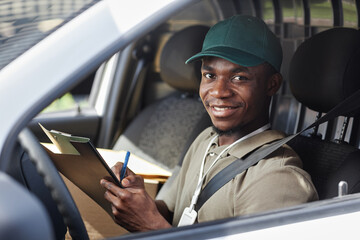 Portrait of young black man writing on clipboard in delivery van and smiling at camera, copy space