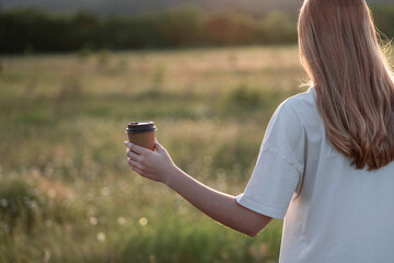 Woman's hand with a paper craft coffee cup in nature in a beautiful sunset light.