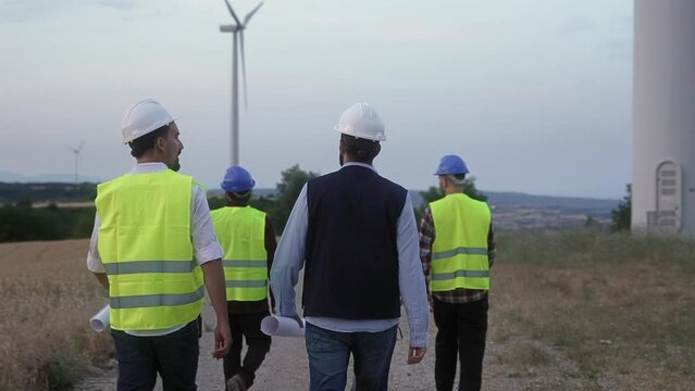 Team of worker specialists and electrical engineers walking around wind turbine farm, doing technical inspection of power station facility at dusk