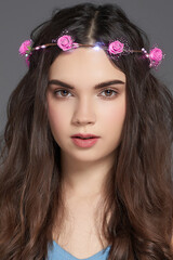 Naklejka premium Cropped close-up shot of a brown-haired young woman with a LED luminous flower wreath on her head. A girl with a pink roses wreath headband is on a gray background. Front view.