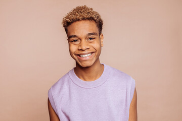 Positive young gay man feels great, wears purple top, broadly smiles and looks at camera stands...
