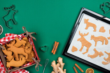 Top view of Christmas gingerbread cookies from raw dough on  baking tray, fresh ginger, cinnamon, deer shaping biscuit,  freshly backed cookies  over green background.Flat lay.