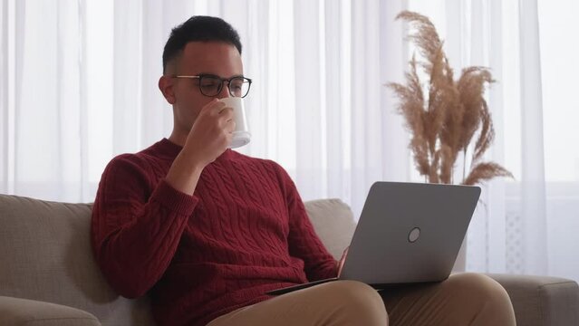 Digital weekend. Home rest. Freelancer leisure. Relaxed man with mug enjoying using notebook watching movie drinking coffee on sofa at modern home interior with free space.