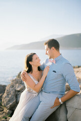 Bride and groom sit hugging on a rocky seashore