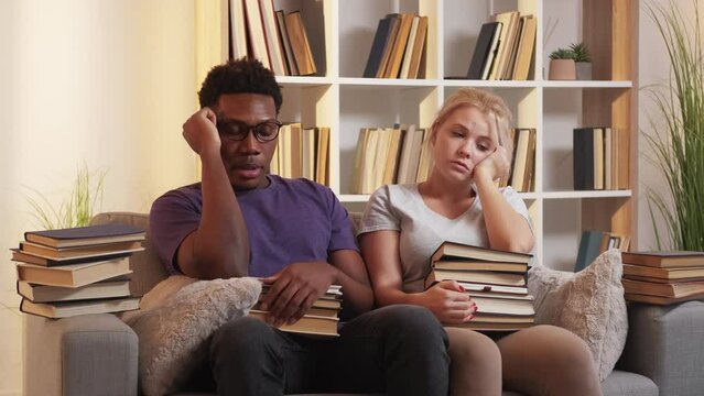 Boring study. Learning fatigue. Exhausting education. Tired lazy two multiracial students sithing on couch with books at home living room interior.