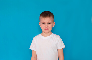 Naklejka premium boy with blond hair, aged five or six, is crying on an isolated blue background. Upbringing, sad, offended child. Close-up of a crying boy, studio shot