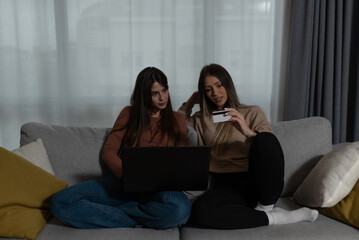 Two young women roommates sitting in the dark at apartment trying to pay electricity bill with credit card online on their laptop computer.