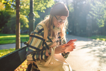 Cheerful young student freelancer woman sit on bench in autumn park outdoors using smartphone at the park.People urban lifestyle concept. 