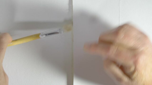 Closeup of a man&rsquo;s hands using a small brush and paste to glue back two sections of wallpaper which have come apart, then pressing the repair area into place. 