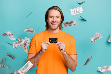 Photo of handsome cheerful man wear orange t-shirt money flying holding credit card isolated turquoise color background