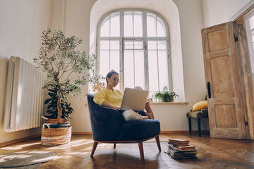 Beautiful young woman using laptop while sitting in a comfortable chair at home
