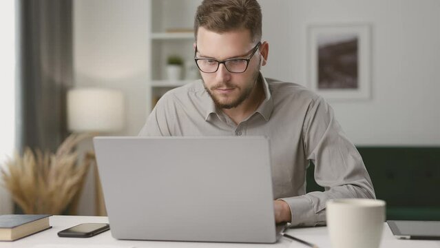 Concentrated young man using laptop, taking notes during remote lecture. Student studying online at home, looking at laptop screen