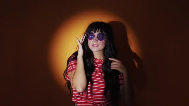 Studio Shot Of Attractive Young Girl Wears Her Round Purple Lens Sunglasses And Poses To The Camera Smiling With Orange Light Background. - Medium Closeup Shot