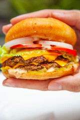 Hands about to lift a queso melted cheese burger with lush green background inside a cafe.