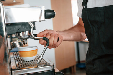 Using coffee machine. Cafe worker in white shirt and black apron is indoors