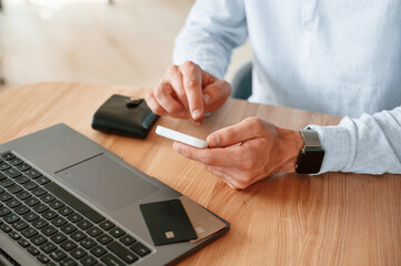 Credit card is on the laptop. Close up view of man that sitting by the table and working by using smartphone