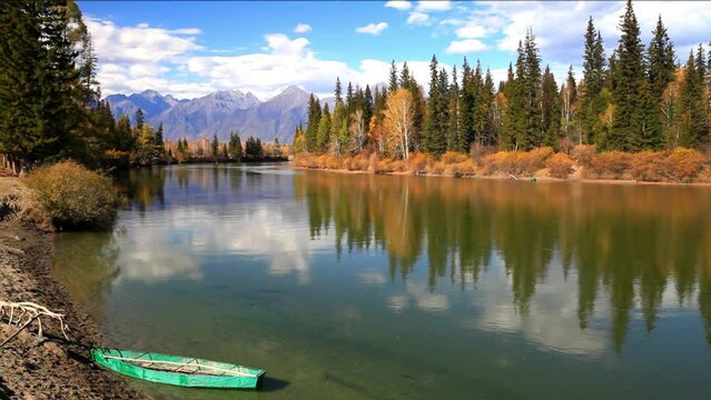 Autumn landscape with yellowed forest on banks of Irkut River and green fishing boat on coast. Mountain range of Eastern Sayan Mountains in distance on sunny September day. Buryatia, Tunka valley 