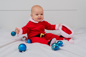 Baby boy dressed as Santa is playing with Christmas decorations on the bed.