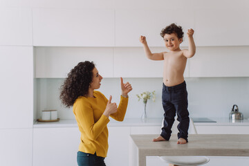Little curly boy shows his biceps, looking at camera standing on table and his mother looking at son gesturing thumbs up, proud of him. Young curly woman satisfied by healthy cheerful son. Family