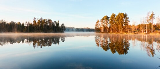 Panoramic view of the forest lake at sunrise. Soft golden sunlight, clear blue sky, reflections on water, fog, frost. Mighty trees. Finland. Nature, seasons, ecology, environment, eco tourism themes - obrazy, fototapety, plakaty