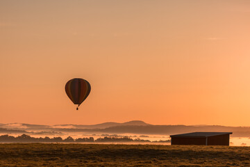 Hot air balloon is basked in a golden glow as it slowly ascends over a fog-filled valley in Orange...
